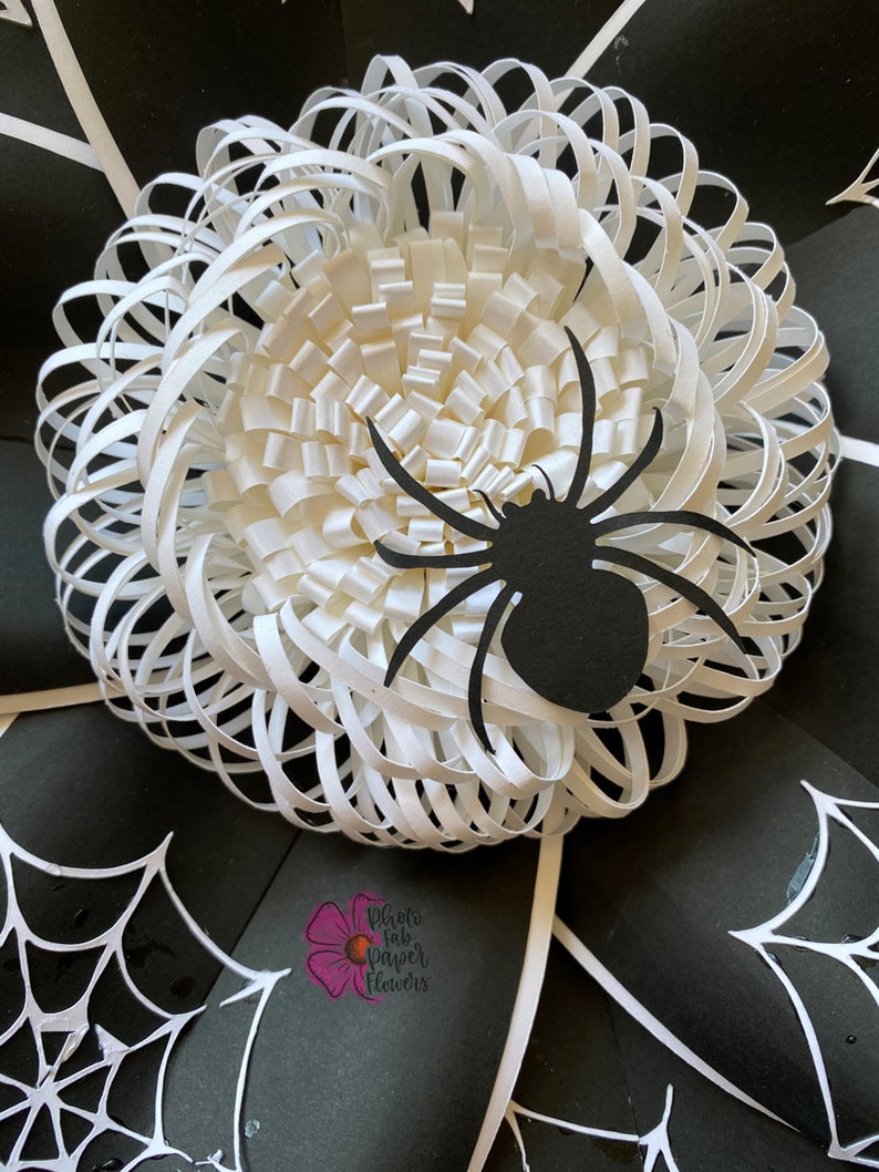 May include: A white paper flower with a black spider on top. The flower is made of many layers of paper, creating a 3D effect. The spider is a simple silhouette with eight legs. The background is black with white spiderwebs.