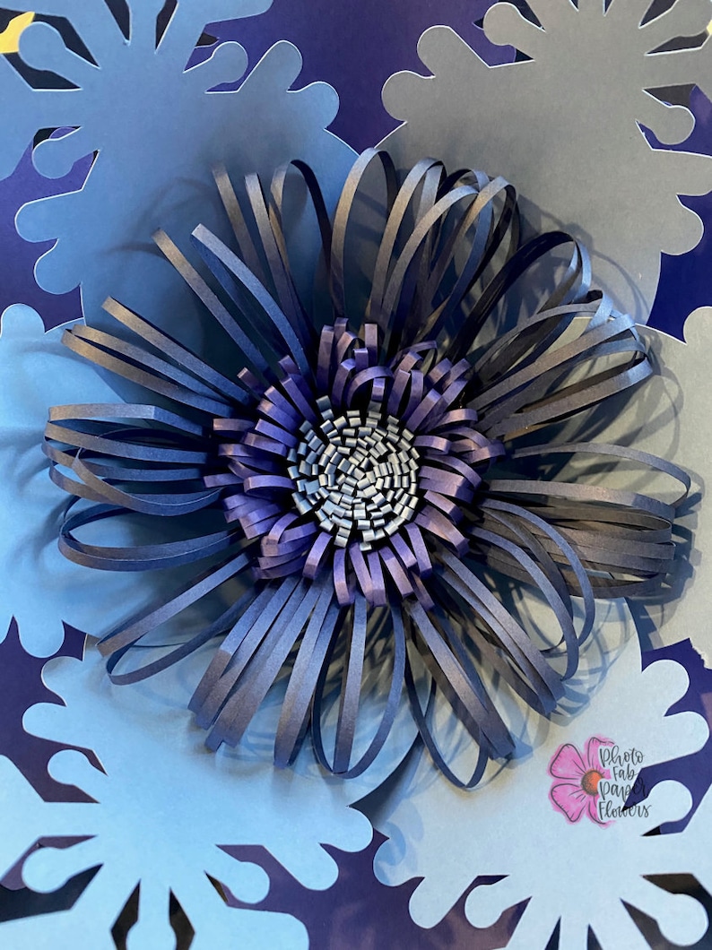May include: A blue paper flower with a layered center, surrounded by blue paper snowflakes. The flower is made of strips of paper that are curled and layered to create a 3D effect. The snowflakes are cut out of paper and have a delicate, intricate design. The image is titled "Photo Fab Paper Flowers".