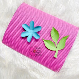 May include: A pink yoga mat with a light blue paper flower and a green paper leaf on top. The text "What to do with paper flowers" is printed on the mat.