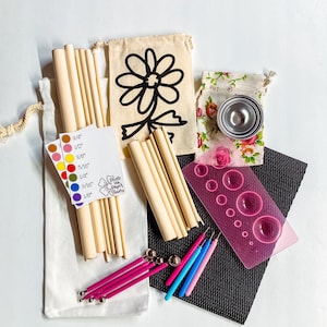 May include: A craft kit for making paper flowers includes a variety of tools, including wooden dowels, a metal cutter, a pink cutting mat, and a set of pink and blue tools. The kit also includes two small fabric bags, one with a black and white daisy design and the other with a floral pattern. The kit includes a guide with different sizes of circles marked in inches.