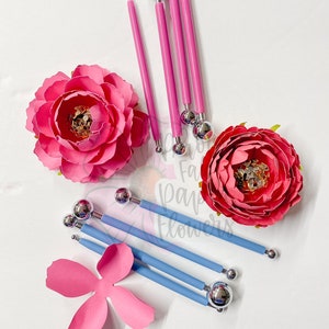 May include: A set of pink and blue paper flower tools with ball-shaped tips. The tools are used to create realistic-looking paper flowers. The tools are arranged on a white surface with pink paper flower petals.