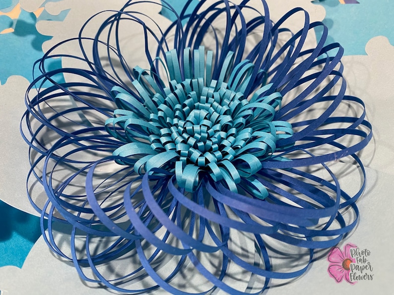 May include: A blue and turquoise paper flower with a layered design. The flower is made of multiple strips of paper that are curled and layered to create a 3D effect.