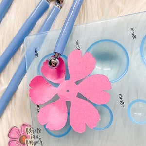 May include: A close-up of a blue plastic flower shaping tool with a pink paper flower on a clear plastic mat with circles labeled in millimeters. The tool has a silver ball on the end. The mat has the text "Photo Fab Paper Flowers" in the lower left corner.