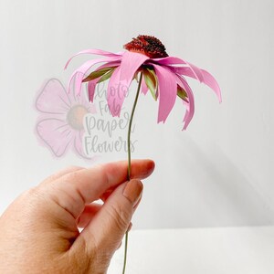 K&ouml;nnte beinhalten: Eine einzelne rosa Papierblume mit brauner Mitte und gr&uuml;nem Stiel. Die Blume wird in einer Hand gehalten.