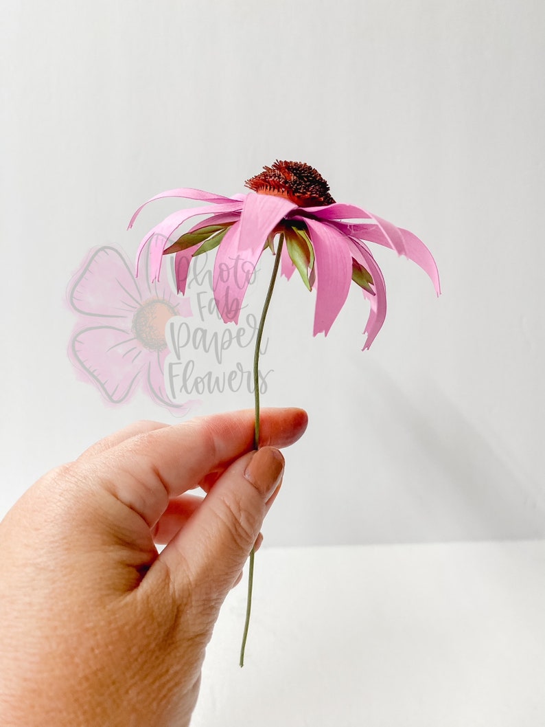 K&ouml;nnte beinhalten: Eine einzelne rosa Papierblume mit brauner Mitte und gr&uuml;nem Stiel. Die Blume wird in einer Hand gehalten.