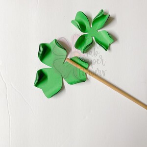 May include: Green paper flower with five petals, partially assembled on a wooden stick. The flower is made of multiple layers of paper.