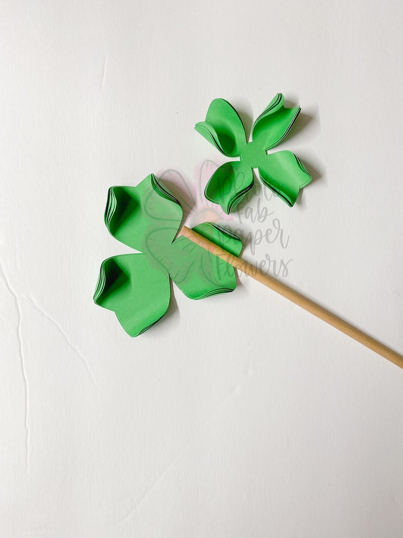 May include: Green paper flower with five petals, partially assembled on a wooden stick. The flower is made of multiple layers of paper.