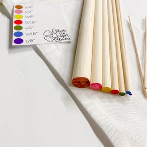 May include: A set of wooden dowels in various sizes, ranging from 1/8 inch to 3/4 inch, with colored tips. The dowels are arranged in a rainbow pattern, with the smallest dowel at the top and the largest at the bottom. The dowels are in a white fabric bag with a tag that reads "Photo Fab Paper Flowers".