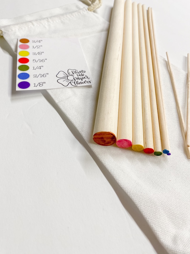 May include: A set of wooden dowels in various sizes, ranging from 1/8 inch to 3/4 inch, with colored tips. The dowels are arranged in a rainbow pattern, with the smallest dowel at the top and the largest at the bottom. The dowels are in a white fabric bag with a tag that reads "Photo Fab Paper Flowers".