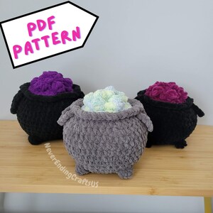 May include: Three crocheted cauldron pillows, one gray, one black, and one purple. Each cauldron has a different colored filling, green, purple, and pink. The pillows are on a wooden surface. The text "PDF PATTERN" is in a pink box in the upper left corner of the image.