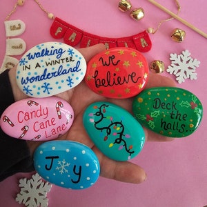 May include: A hand holding seven painted stones with Christmas-themed designs and sayings. The stones are painted in various colours, including red, green, blue, pink, and white. The sayings on the stones include "walking in a winter wonderland", "we believe", "candy cane lane", "deck the halls", and "joy".