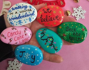 May include: A hand holding seven painted rocks with Christmas-themed designs and sayings. The rocks are painted in various colors, including red, green, blue, pink, and white. The sayings on the rocks include "walking in a winter wonderland", "we believe", "candy cane lane", "deck the halls", and "joy".
