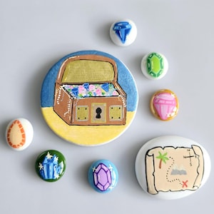 May include: A collection of hand-painted stones. One depicts a treasure chest filled with gems, others show individual gemstones in various colours, and one has a treasure map design. The chest is brown and gold.