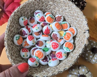 May include: A woven basket filled with white, oval-shaped stones decorated with fall-themed designs. The designs include pumpkins, mushrooms, and autumn leaves in orange, red, and brown. A yellow pumpkin and fall foliage are in the background.