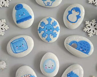 May include: A collection of ten smooth white stones painted with blue winter-themed illustrations including a beanie, mittens, a penguin, a snowflake, an ice cube, an igloo, a snowball, a mug of cocoa, and a pine tree, surrounded by felt balls.