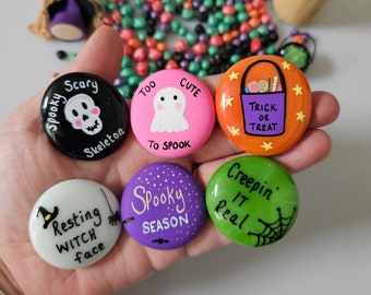 May include: Six round buttons featuring Halloween designs. One black button says Spooky Scary Skeleton, a pink one says Too Cute To Spook, an orange one says Trick or Treat, a white one says Resting Witch face, a purple one says Spooky Season, and a green one says Creepin IT Real.