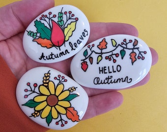 May include: Three white, oval-shaped stones with hand-painted autumn-themed designs. One stone says "Autumn leaves" with a leaf design. Another says "Hello Autumn" with leaf and berry designs. The third stone features a sunflower design.