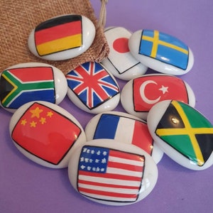 May include: A set of ten oval-shaped ceramic stones, each painted with a different country's flag. The flags include Germany, Japan, Sweden, South Africa, United Kingdom, Turkey, China, France, the United States, and Jamaica.