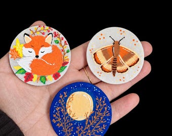 May include: Three circular, hand-painted wooden discs featuring distinct designs: a sleeping orange fox surrounded by autumn leaves, a brown and orange moth with spotted wings, and a yellow full moon set against a dark blue starry night sky.