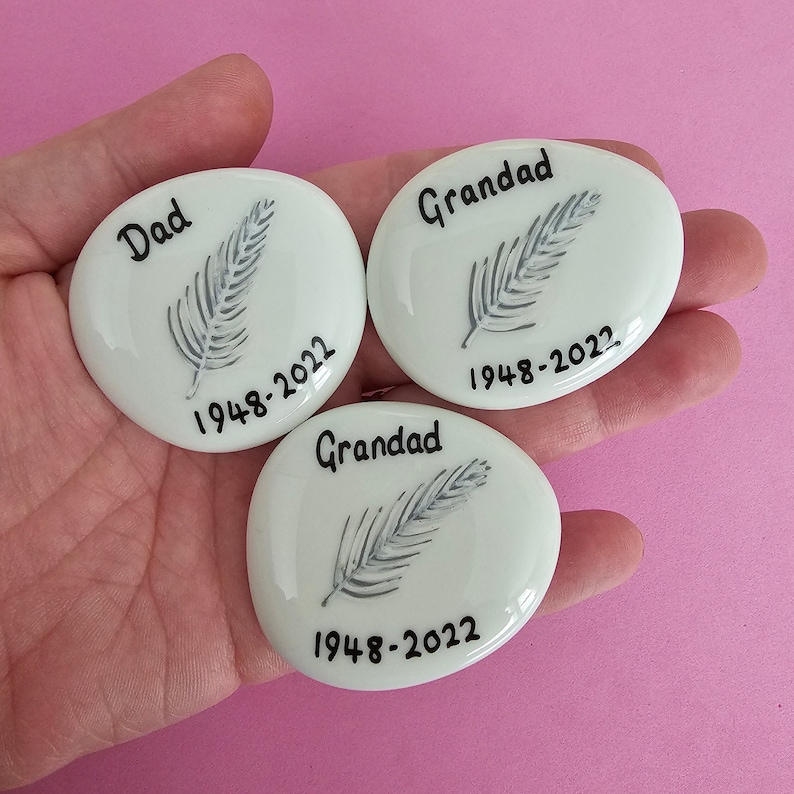 white oval-shaped pebbles with black lettering and a grey feather design. The pebbles can be personalised with a name on top and or dates on the bottom