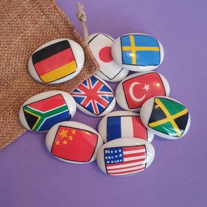 May include: A set of nine oval-shaped ceramic tiles with flags of different countries painted on them. The flags include Germany, Japan, Sweden, the United Kingdom, Turkey, Jamaica, South Africa, China, France, and the United States.
