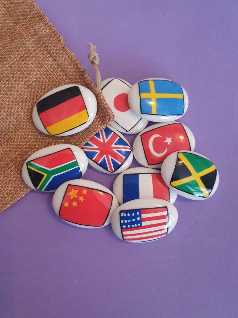 May include: A set of nine oval-shaped ceramic tiles with flags of different countries painted on them. The flags include Germany, Japan, Sweden, the United Kingdom, Turkey, Jamaica, South Africa, China, France, and the United States.
