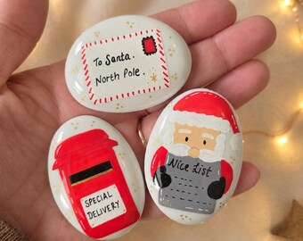 May include: Three smooth, white, oval-shaped stones painted with Christmas themes. One stone features an envelope addressed to Santa, North Pole. Another shows a red post box labeled Special Delivery. The third depicts Santa holding a Nice List.