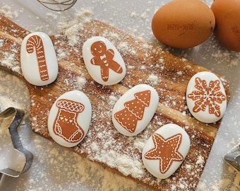 May include: Six white painted stones with brown gingerbread designs. The designs include a candy cane, gingerbread man, Christmas tree, stocking, snowflake, and a star. The stones are on a wooden cutting board with flour sprinkled on it.