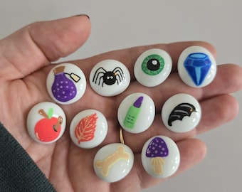 May include: A collection of ten small, round, white buttons featuring various colorful illustrations including a purple potion bottle, a black spider, a green eye, a blue diamond, a red apple with a worm, an orange leaf, a green finger, a black bat wing, a tan bone, and a purple mushroom.