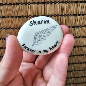 May include: White ceramic stone with a black feather design and the text "Sharon forever in my heart".