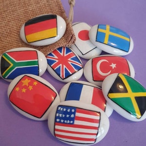 May include: A set of ten oval-shaped, white ceramic stones with flags of different countries painted on them. The flags include Germany, Japan, Sweden, South Africa, United Kingdom, Turkey, China, France, the United States, and Jamaica.
