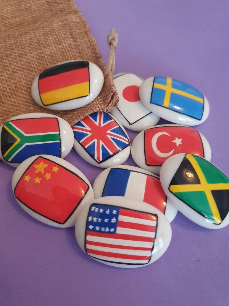 May include: A set of ten oval-shaped, white ceramic stones with flags of different countries painted on them. The flags include Germany, Japan, Sweden, South Africa, United Kingdom, Turkey, China, France, the United States, and Jamaica.