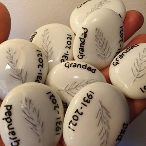 May include: White ceramic stones with a gray feather design and black text. The text includes "Grandad" and dates.