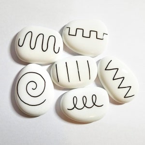 May include: Six white smooth stones with black line designs. The designs include a wavy line, a spiral, a zig-zag, a series of vertical lines, a series of horizontal lines, and a series of steps.