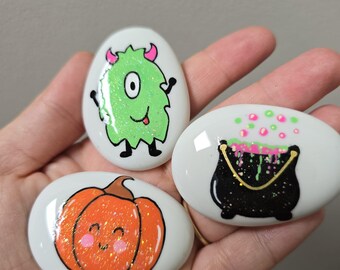 May include: Three smooth, white, oval-shaped stones featuring hand-painted designs: a glittery lime green monster with pink horns, a bright orange pumpkin with a smiling face, and a black cauldron with bubbling pink and green liquid.