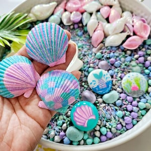 May include: A hand holding three iridescent seashells with pink and blue glitter. The shells are on top of a bowl filled with purple, blue, and white rice, and small decorative rocks with mermaid and sea life designs.
