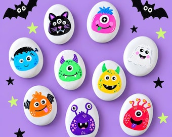 May include: A collection of eight white oval stones painted with colorful monster faces, including a black cat, pink cyclops, blue Frankenstein-style creature, green monster, yellow horned beast, ghost, orange bat-like creature, purple alien, and red monster.