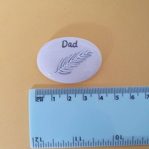 May include: White ceramic stone with a feather design and the word "Dad" written on it. The stone is about 2 inches in diameter.