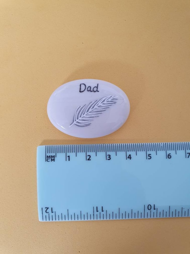 May include: White ceramic stone with a feather design and the word "Dad" written on it. The stone is about 2 inches in diameter.
