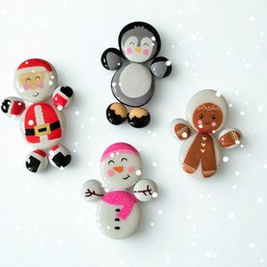 May include: Four painted stone Christmas figures: Santa Claus, a penguin, a snowman with a pink scarf, and a gingerbread person. Each figure has a unique design and colour scheme, set against a white background with decorative snow.