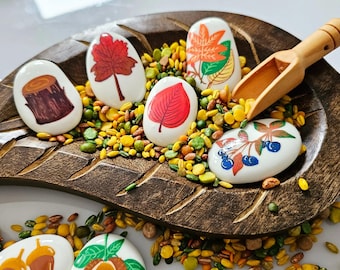 May include: A wooden tray shaped like a leaf filled with small yellow and green beans.  There are six white painted stones with different autumn-themed images on them, including a tree stump, a leaf, a tree, and berries.