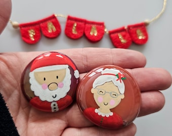 May include: Two painted ornaments, one depicting Santa Claus with a white beard and red hat, and the other depicting a woman with white hair and a red hat, both with smiling faces.