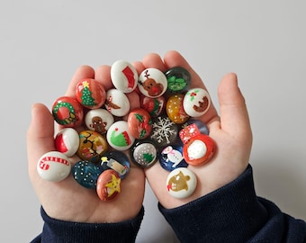 May include: A collection of Christmas-themed decorative stones, each with unique hand-painted designs. The stones feature festive images like Christmas trees, reindeer, snowflakes, and gingerbread men. The stones are held in open hands against a plain background.