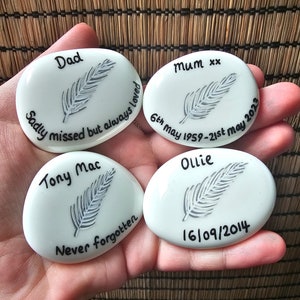 May include: Four white oval-shaped stones with black lettering and a gray feather design. The stones read "Dad Sadly missed but always loved", "Mum xx 6th May 1959-21st May 2023", "Tony Mac Never forgotten", and "Ollie 16/09/2014".