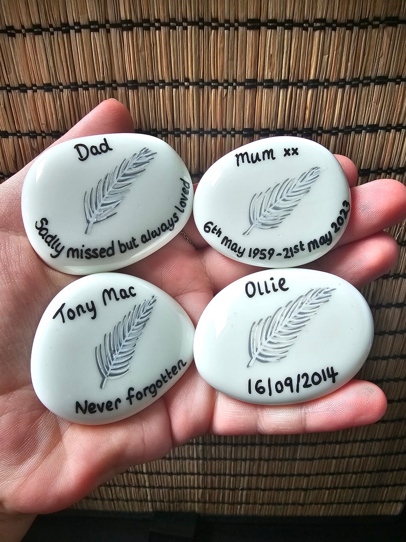 May include: Four white oval-shaped stones with black lettering and a gray feather design. The stones read "Dad Sadly missed but always loved", "Mum xx 6th May 1959-21st May 2023", "Tony Mac Never forgotten", and "Ollie 16/09/2014".