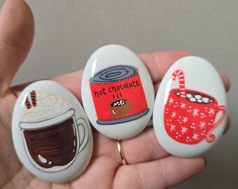 May include: Three painted stones with hot beverage designs. One stone features a coffee cup with whipped cream, another a red can of hot chocolate, and the third a red mug with a candy cane and snowflakes. The stones are white with painted illustrations.