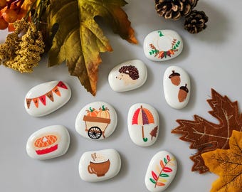 May include: Nine painted white rocks with autumn-themed designs. The designs include a pumpkin patch, a hedgehog, a pie, a wreath, an umbrella, a cup of coffee, a fall leaf, acorns, and a banner that says "Autumn".