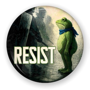May include: A round button with a green frog wearing a blue scarf, standing in front of a group of silhouetted figures. The word "RESIST" is printed in large, white letters.