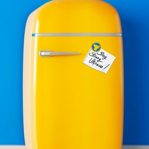 Pu&ograve; includere: Un frigorifero vintage giallo con un biglietto bianco che dice "Stay Strong Ukraine!" attaccato alla porta con un magnete a forma di uccello blu e giallo.