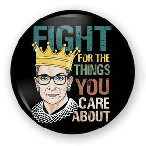 May include: A round black pinback button featuring an illustration of a person wearing a gold crown, black glasses, teal earrings, and a white lace collar. The text reads FIGHT FOR THE THINGS YOU CARE ABOUT in distressed teal, yellow, and tan.